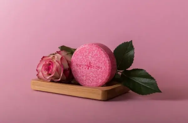 Produktbild pinkes Happy Soaps Shampoo Rose auf einer kleinen Holzunterlage mit einer pinken Rose daneben. Pinker Bild Hintergrund.